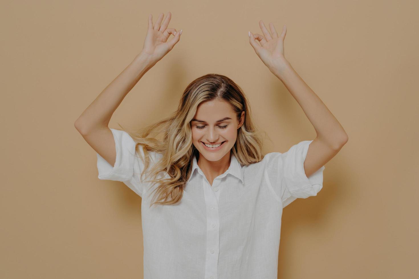 encantadora mujer despreocupada con camisa blanca y cabello rubio disfrutando de un buen día, manteniendo los ojos cerrados y levantando los brazos foto