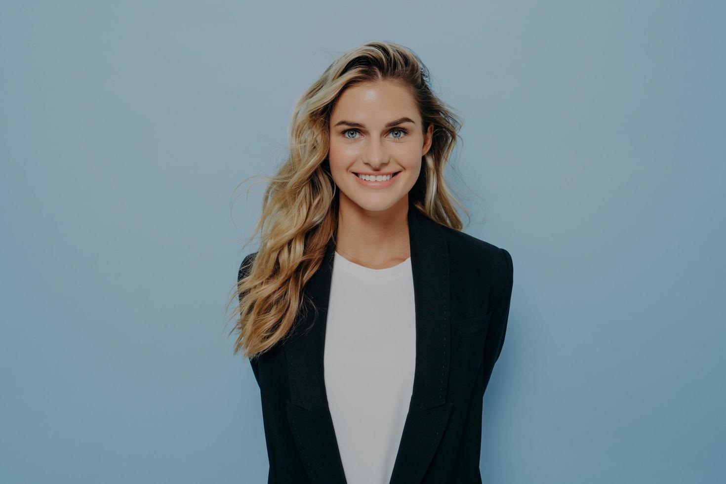 Vivacious blond woman in stylish blazer with happy beaming smile standing with hands behind her back photo