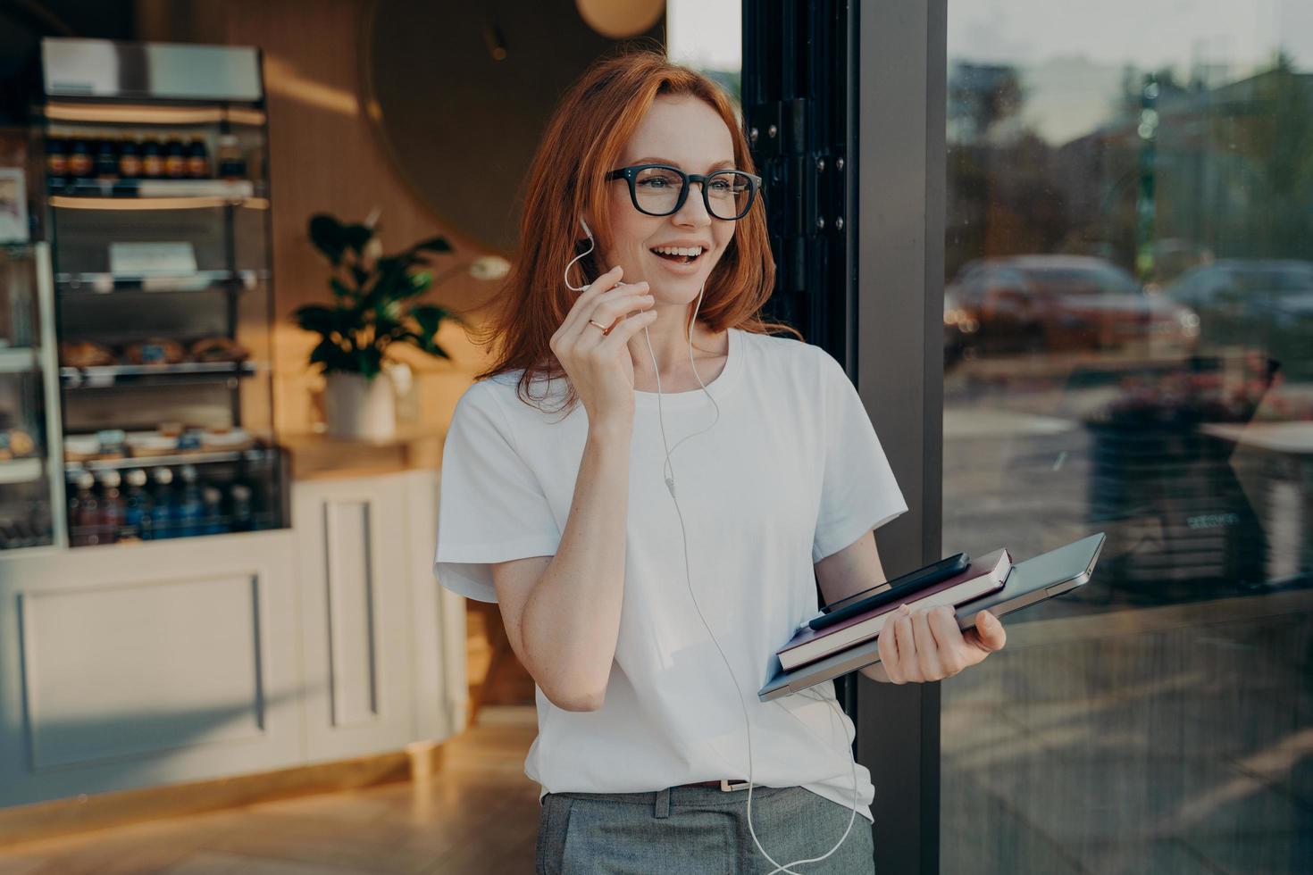 Happy red-haired businesswoman with laptop in hand having call on mobile phone, leaving coffee shop photo