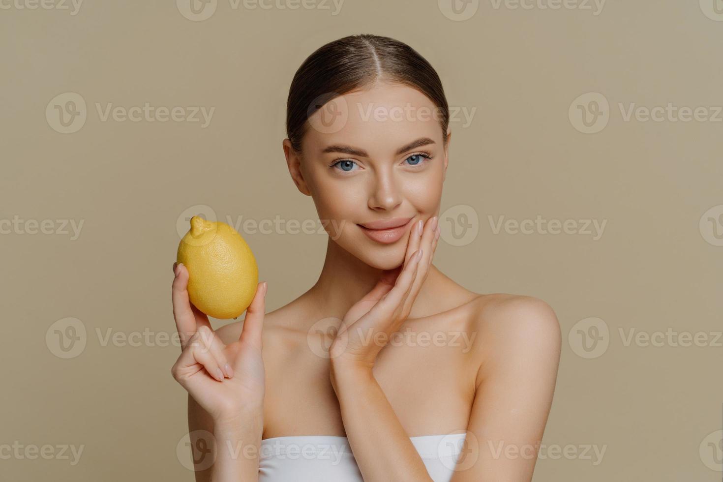 retrato de una hermosa morena europea sostiene limón amarillo fresco para hacer poses de máscara de crema natural envuelta en una toalla aislada sobre fondo marrón. concepto de higiene y belleza para el cuidado de la piel foto