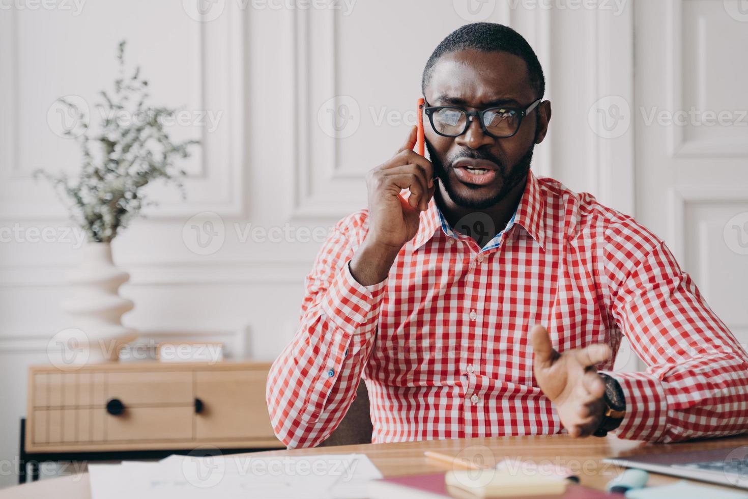 Dissatisfied African American businessman emotionally speaking on smartphone while sitting at desk photo