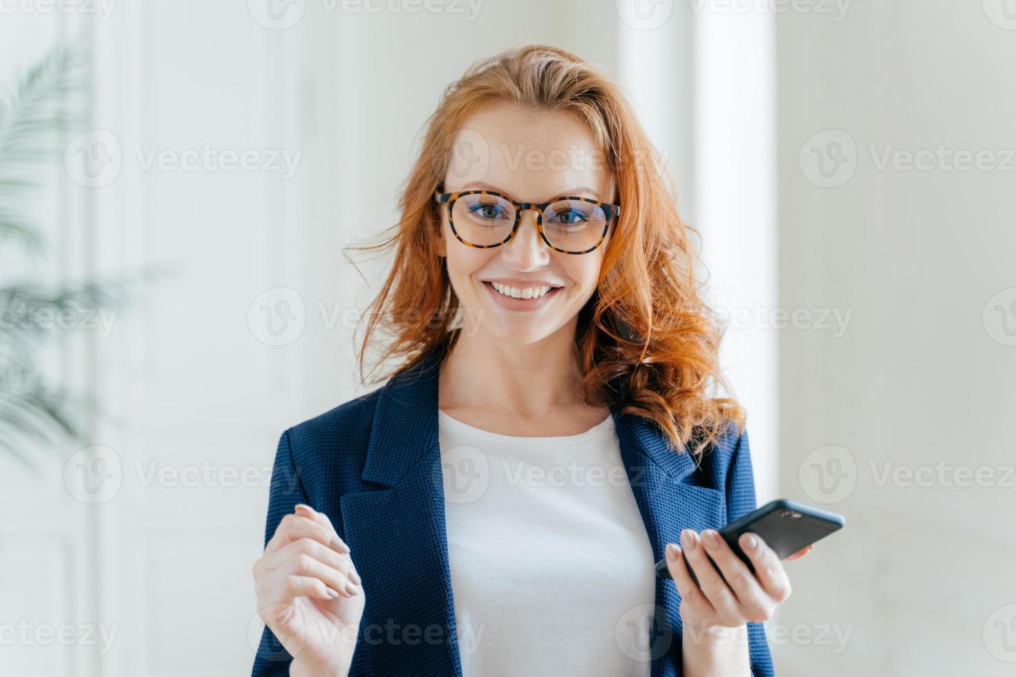 Photo of happy female leader with ginger hair, wears spectacles, updates app on modern cell phone, wears elegant clothes, poses in modern office, reads incoming message from business partner