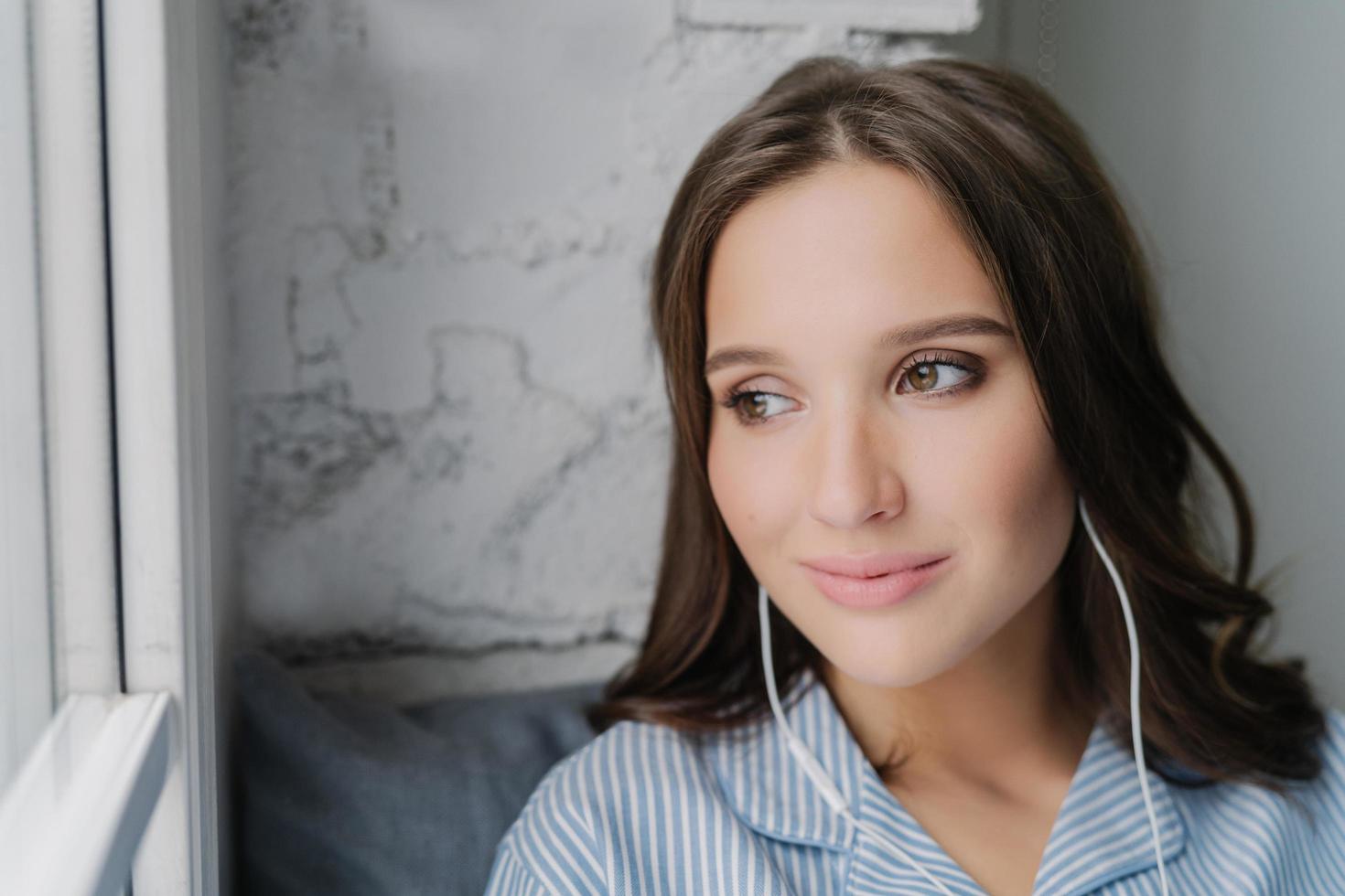 Headshot of attractive female meloman enjoys music in earphones with high volume, likes peaceful atmosphere, looks thoughtfully at window, listens favourite song. People, rest, technology concept photo