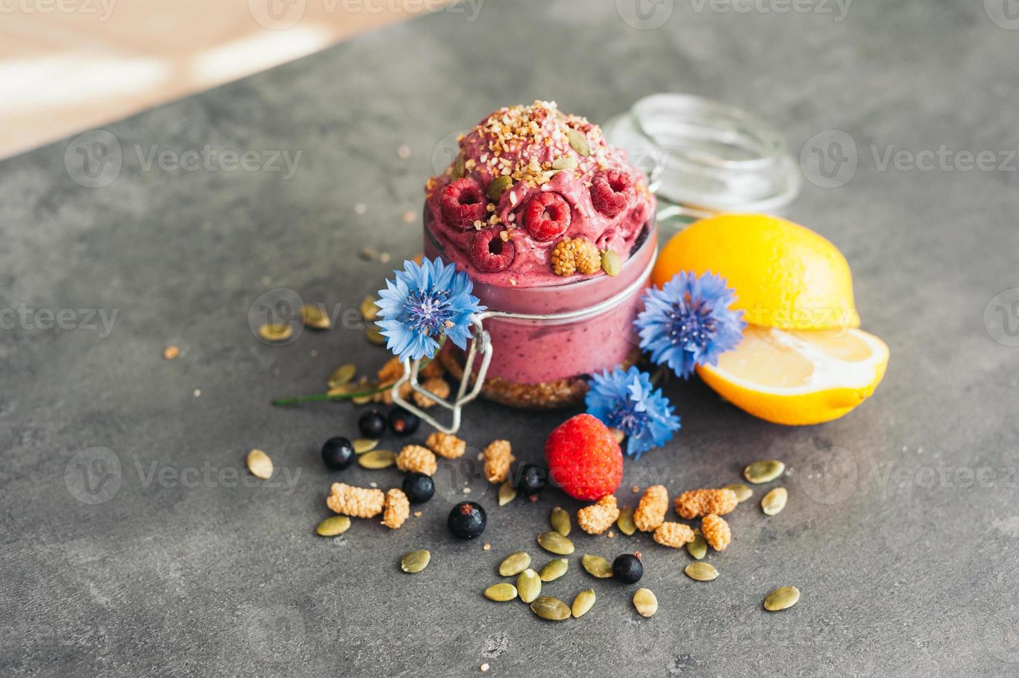 delicioso helado de frambuesa con semillas de calabaza y semillas de cáñamo, decorado con flores azules, grosella negra, limón en rodajas sobre una superficie gris. sabroso postre foto