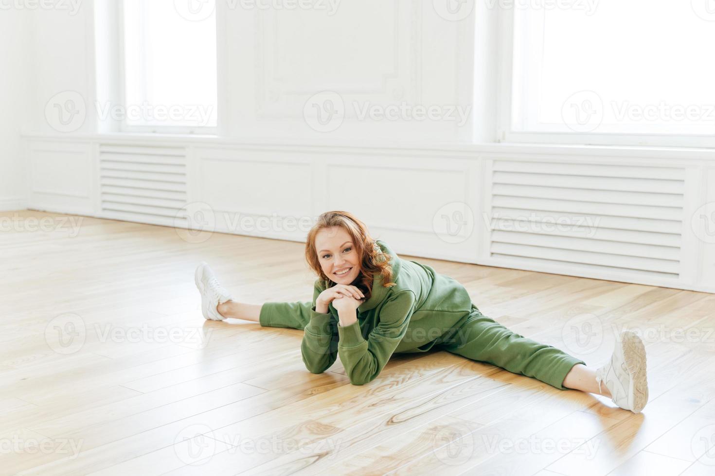 Glad refreshed sporty red haired woman has work out in empty hall, demonstrates flexibility exercise, keeps hands under chin, dressed in green tracksuit and sneakers. Yoga and fitness concept. photo