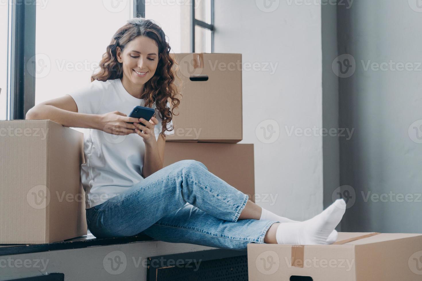 feliz mujer española está sentada en el alféizar de la ventana enviando mensajes de texto por teléfono y desempacando cajas. foto
