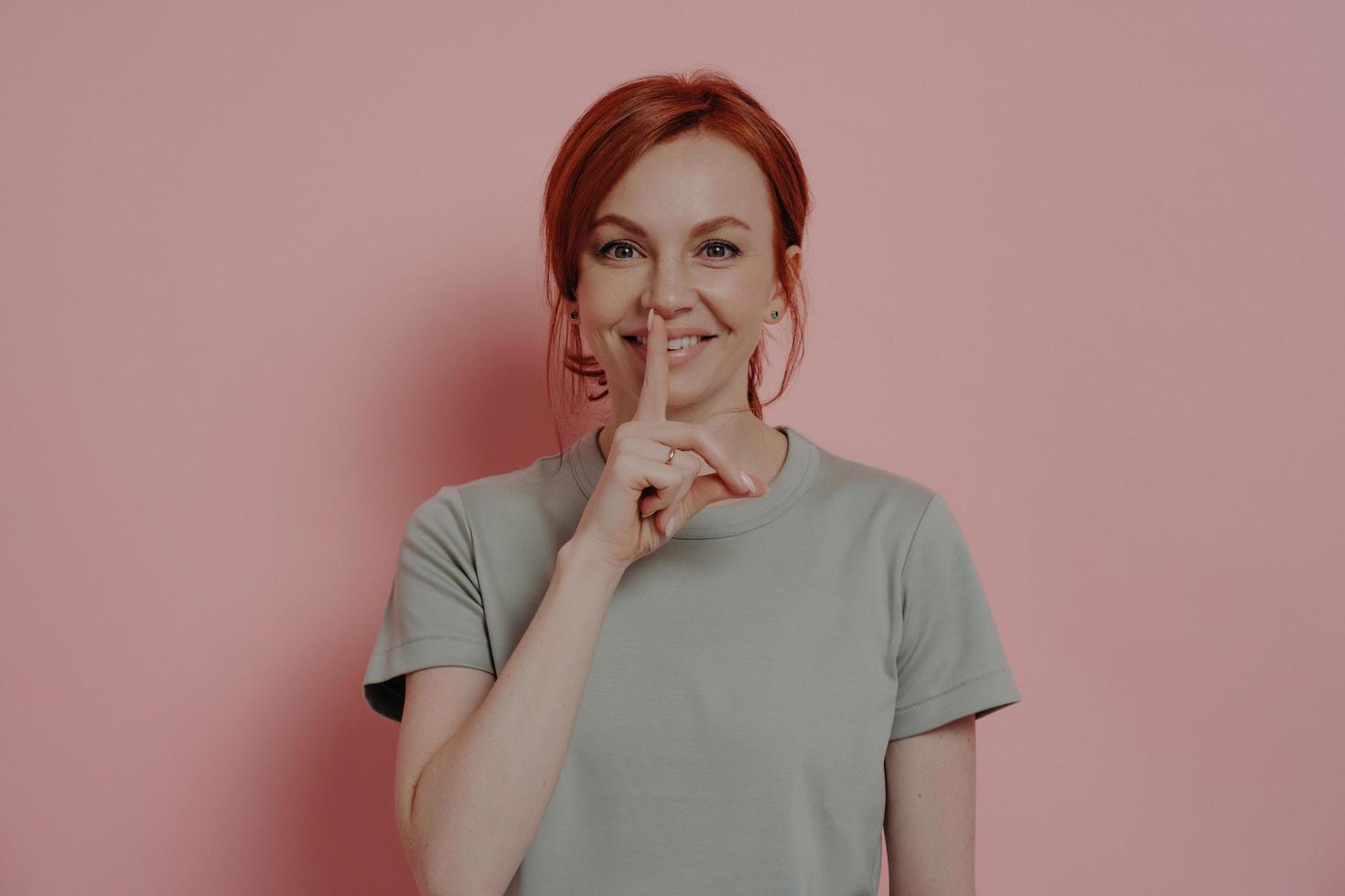 Positive young ginger girl making shush gesture, keeping forefinger over lips, isolated on pink photo