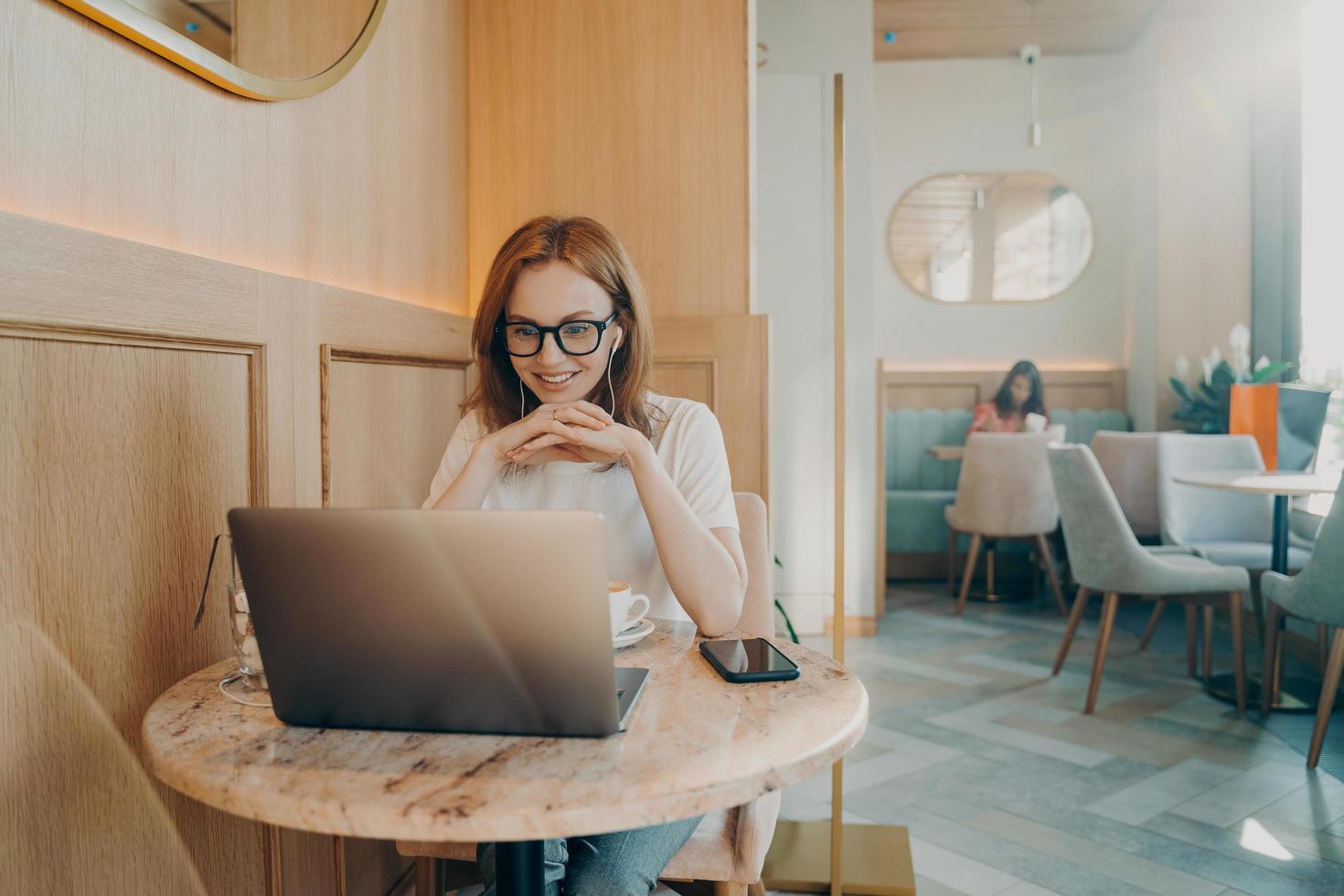 Happy redhaired woman has distance job works on laptop computer in cozy cafe photo