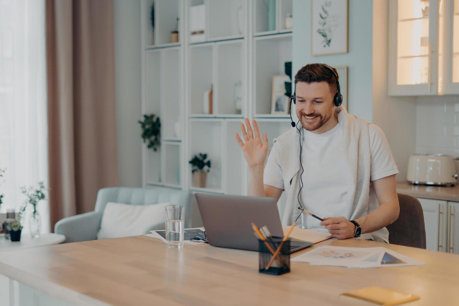 Happy male freelancer using headset and working at home during quarantine while looking at laptop screen photo