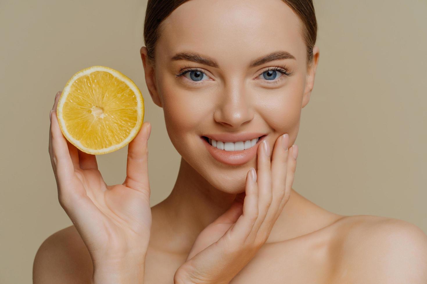Close up shot of beautiful tender woman smiles gently has healthy well groomed skin holds slice of lemon uses citrus fruit for beauty and health stands shirtless isolated over brown background photo