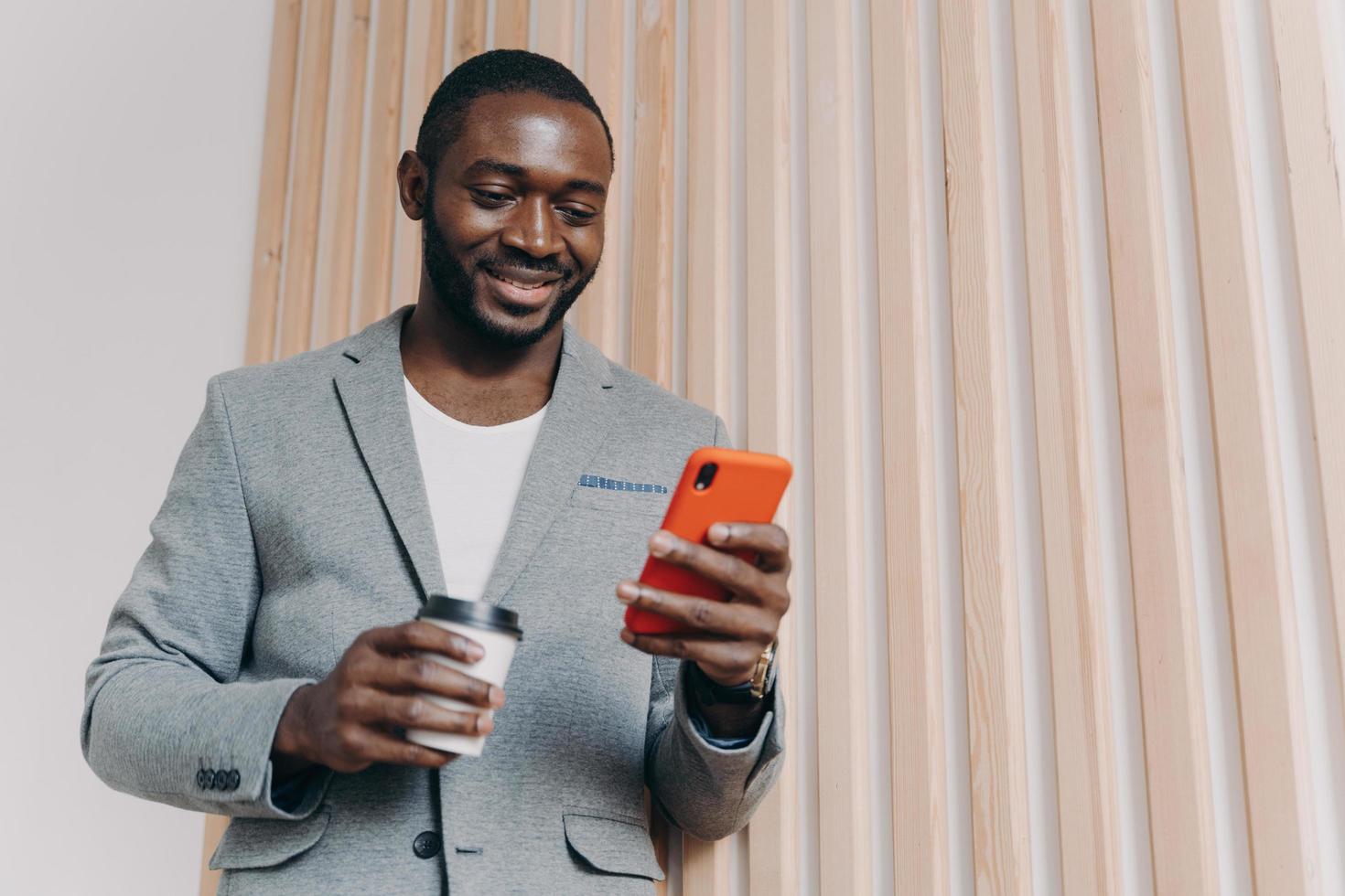 disfrutando del descanso para tomar café en el trabajo. joven trabajador de oficina africano positivo en traje usando teléfono móvil foto