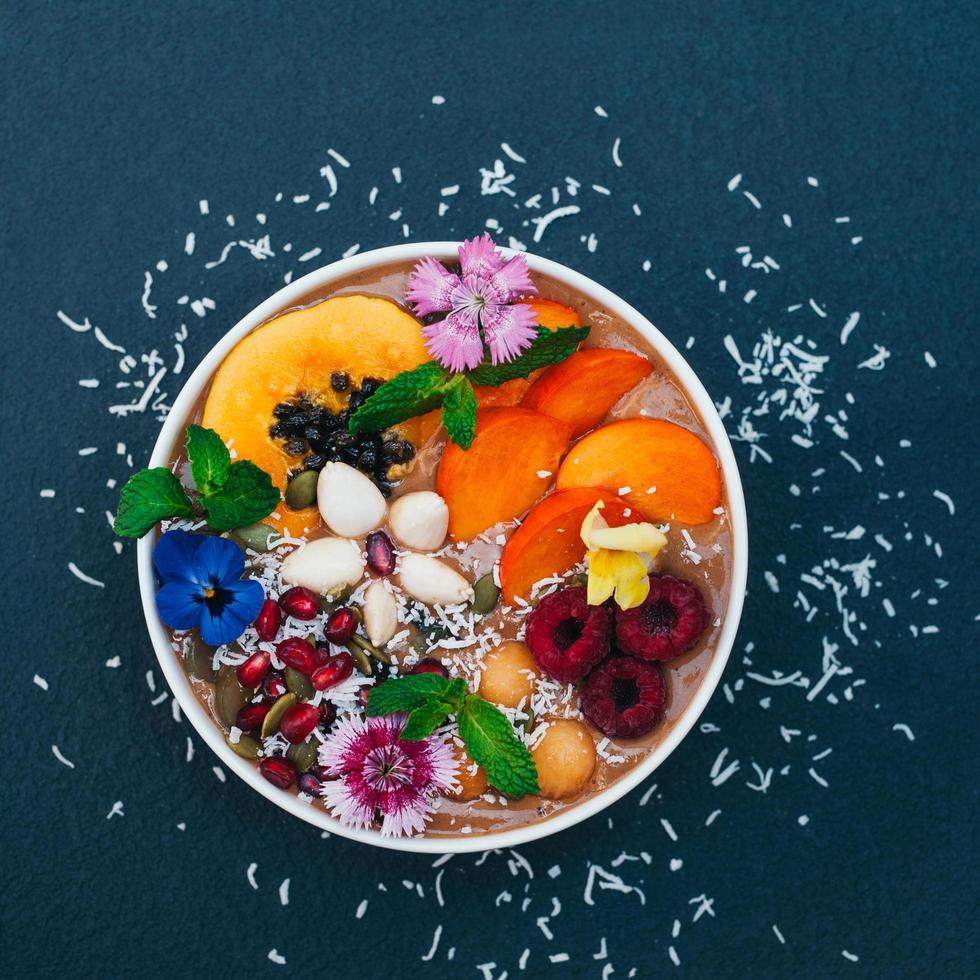 Isolated shot of bowl with tropical fruit decorated with flowers, mint, coconut flakes isolated over dark blue background. Fresh persimone, raspberry and almond. photo