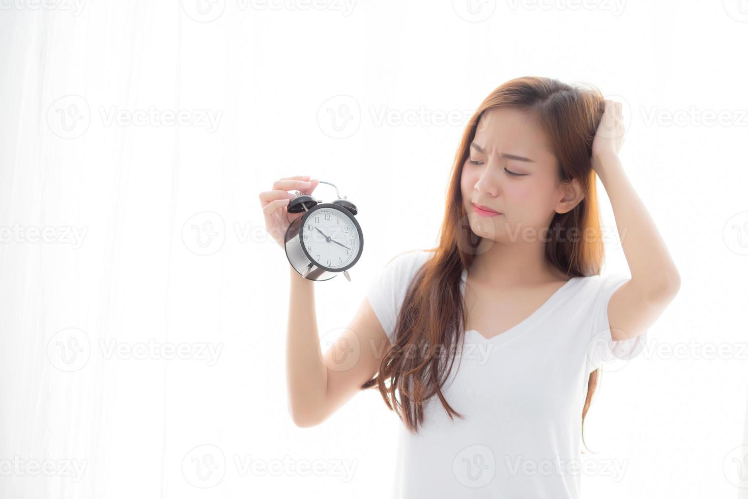 Beautiful young asian woman wake up in morning annoyed alarm clock holding hand, girl standing hurry wake late with appointment with curtain background on bedroom, lifestyle concept. photo