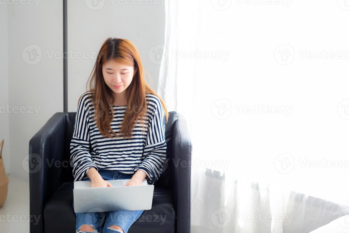 hermoso retrato de una joven asiática que usa una computadora portátil para el ocio en una silla en la sala de estar, una chica que trabaja en línea con un portátil independiente, concepto de negocio de comunicación. foto