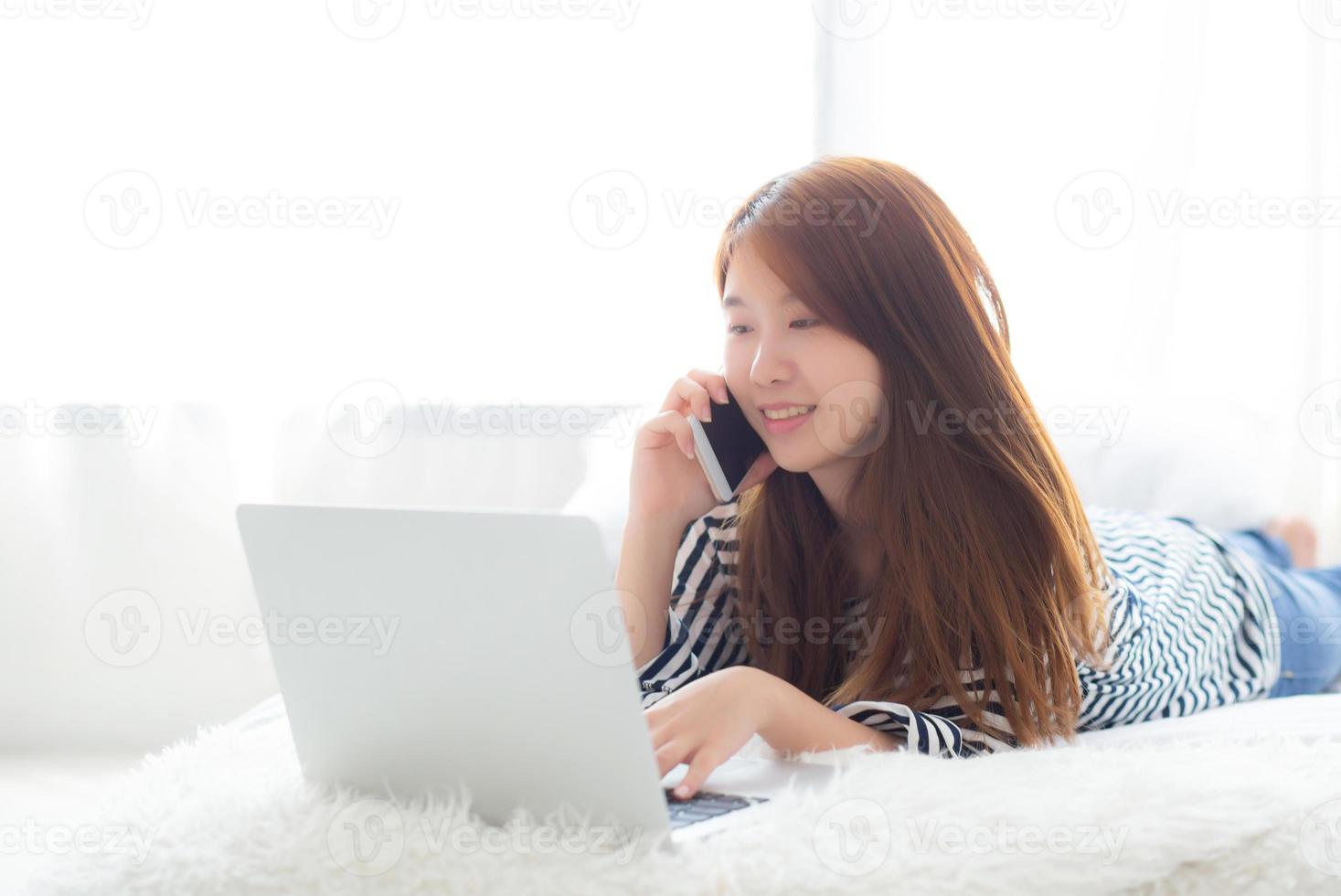 hermosa mujer joven asiática acostada en la cama usando una computadora portátil y hablando de un teléfono inteligente móvil en el dormitorio para el ocio y la relajación, independiente con un cuaderno de trabajo para niñas, concepto de comunicación. foto