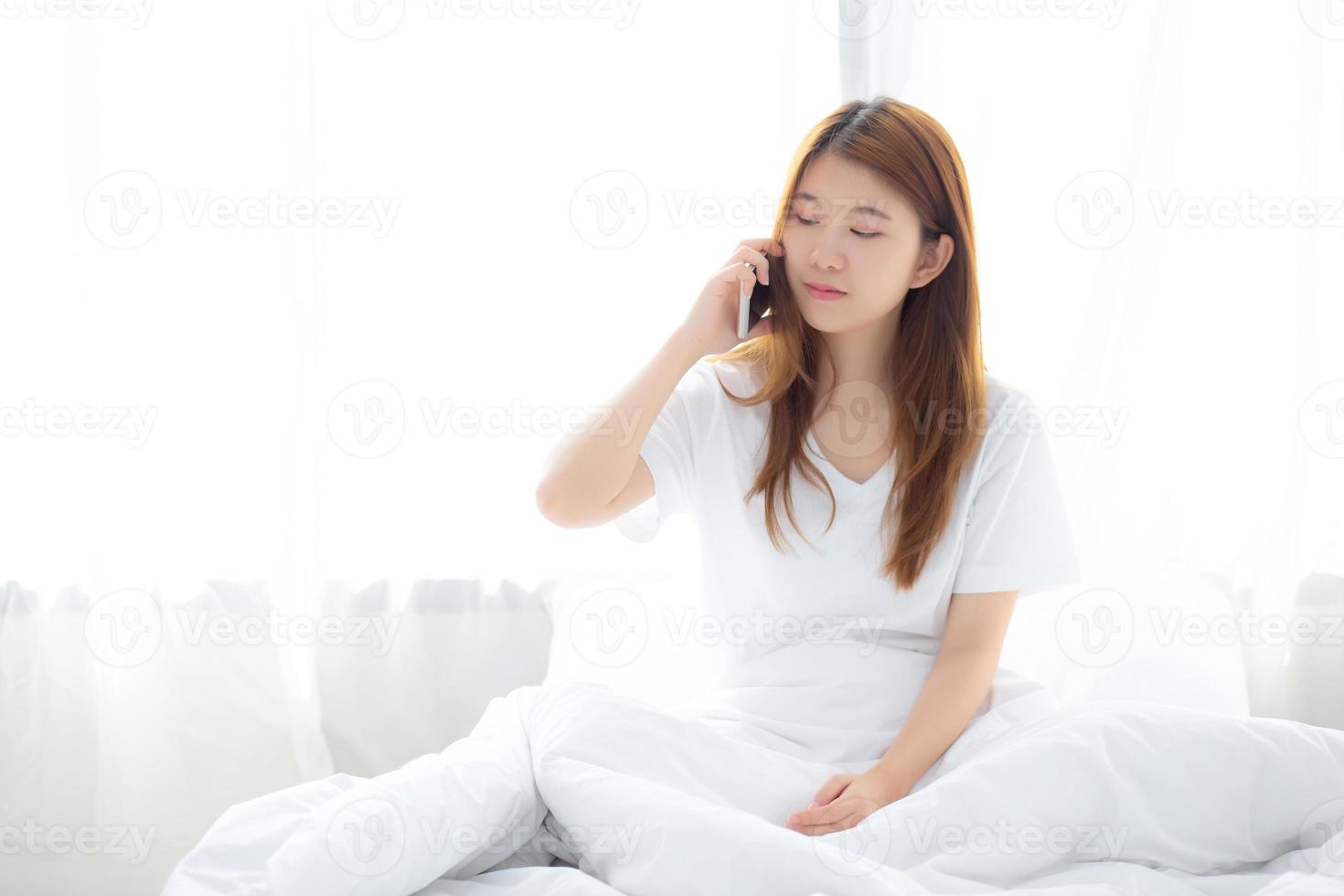 hermosa joven asiática sonriendo sentada y relajándose en la cama por la mañana, chica usando un teléfono móvil inteligente hablando disfrutando en el dormitorio, concepto de comunicación. foto