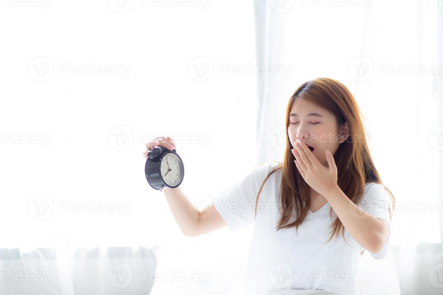 Beautiful young asian woman yawn wake up in morning annoyed alarm clock holding hand, standing hurry wake late with appointment with curtain background on bedroom, lifestyle concept. photo