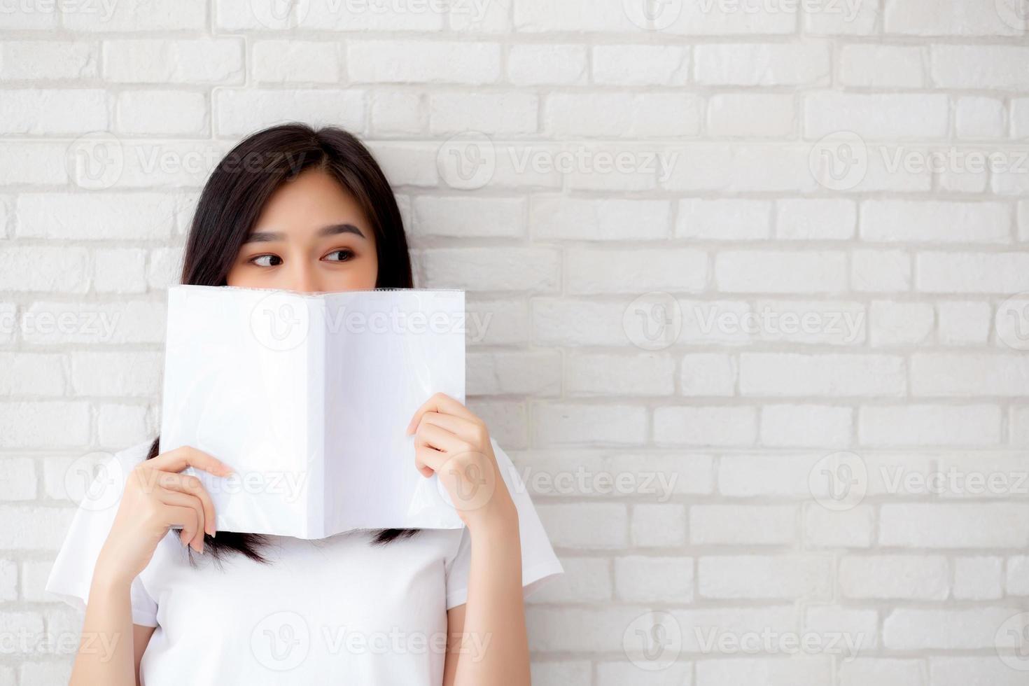 beautiful portrait young asian woman happy hiding behind open the book with cement or concrete background, girl standing reading for learning, education and knowledge concept. photo