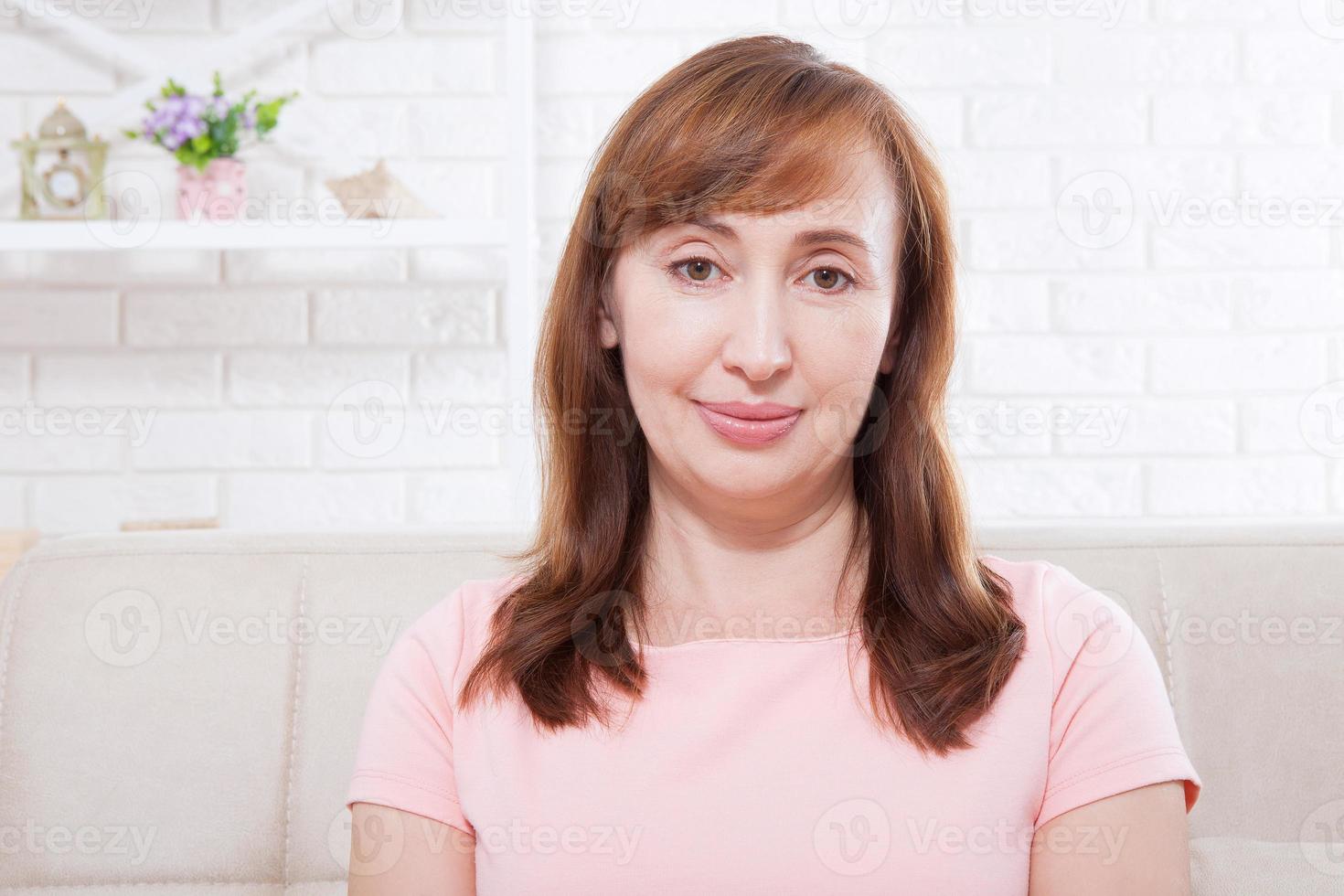 Smiling Beautiful middle-aged brunette woman sitting on sofa at home looking at the camera. Face with wrinkles. Menopause. Copy space and mock up photo