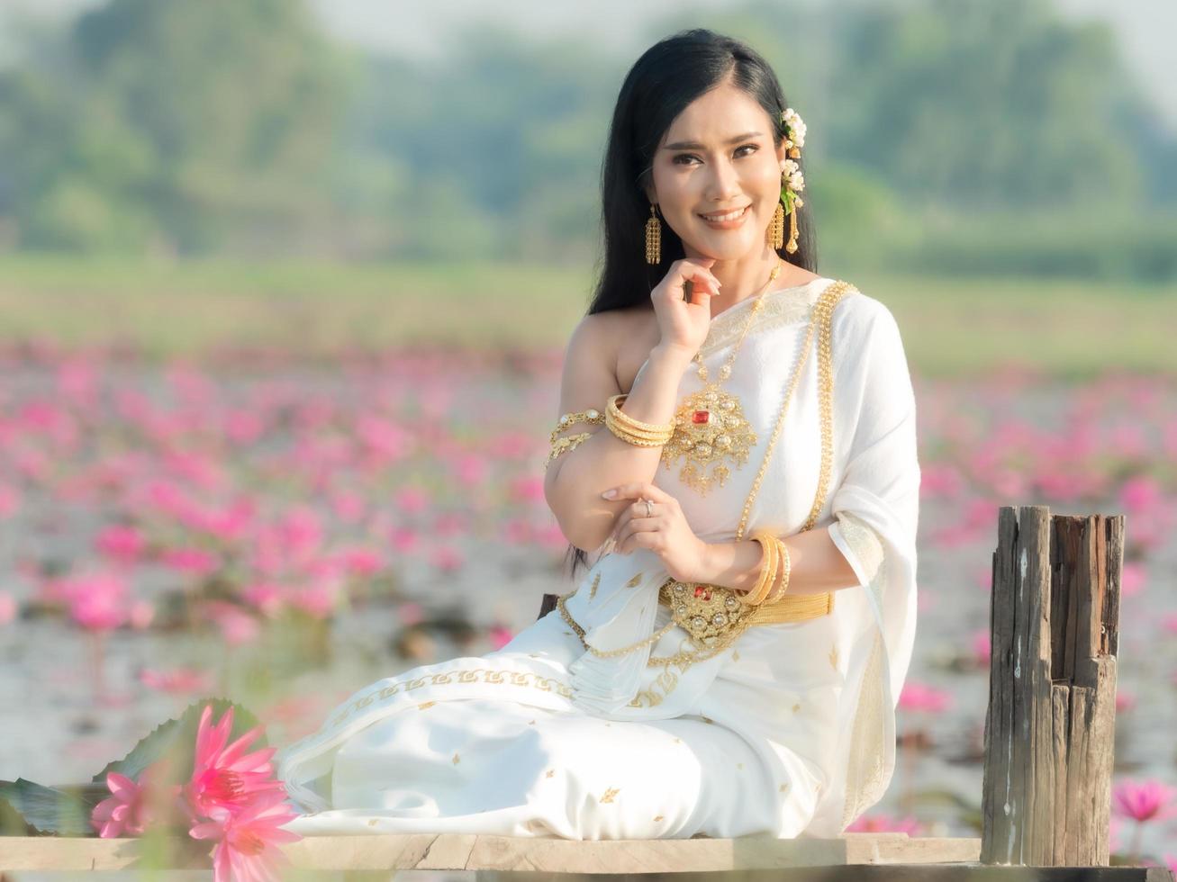 una elegante mujer tailandesa con ropa tradicional tailandesa adornada con adornos dorados, sentada en un puente de madera en un campo de loto foto