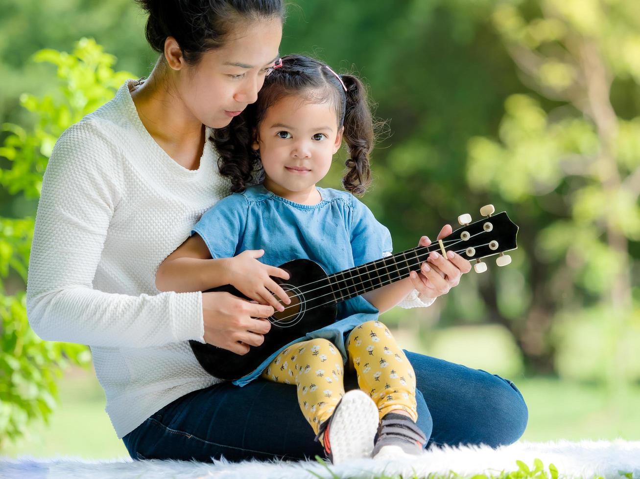 una niña asiática se sienta en la alfombra con su madre y su madre le enseña a tocar el ukelele foto