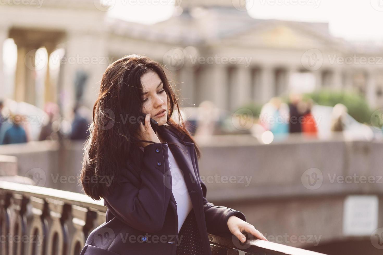 mujer con cabello largo hablando por teléfono. otoño o invierno, chica al aire libre. hermosa morena inteligente en calle de st. petersburgo en el centro de la ciudad cerca del puente foto