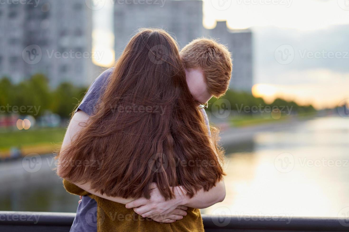 Girl with long thick dark hear embracing redhead boy on bridge, teen love at the sunset photo