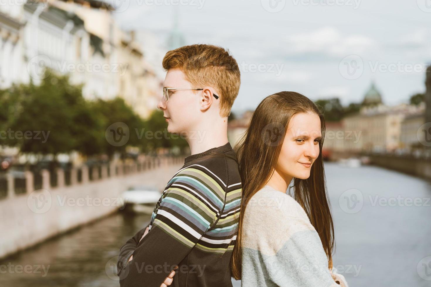 retrato de pareja feliz, de pie espalda con espalda en el centro de la ciudad, hombre pelirrojo con anteojos, mujer hermosa con cabello largo y oscuro foto
