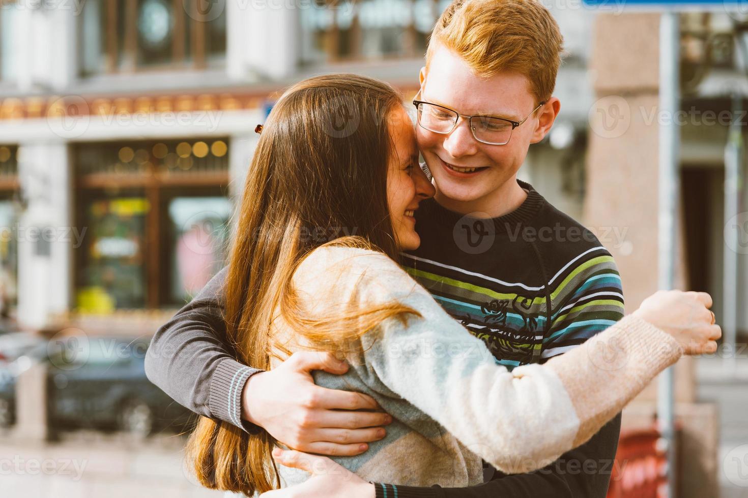 A red-haired young man embraces young woman with long dark hair, they laugh and fool around, standing on a bridge in the center of the city. Concept of first teenage love, serious relationship photo