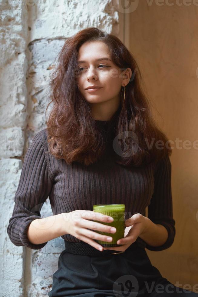 hermosa chica seria, elegante y elegante está sentada en un café y bebiendo un batido verde saludable o vegano con leche. encantadora mujer pensativa con largo cabello castaño oscuro. foto