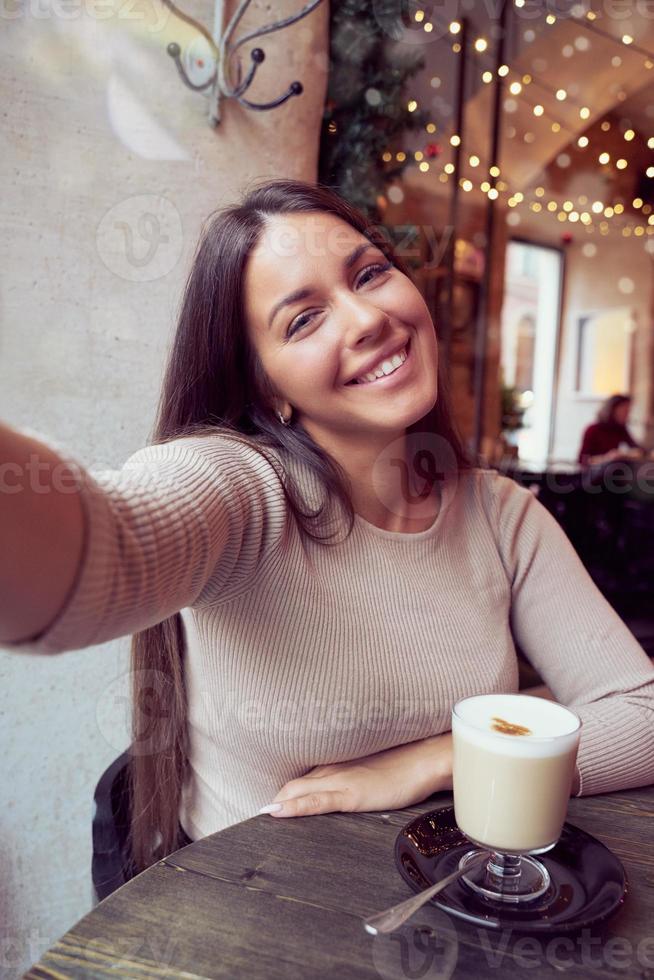 hermosa niña feliz tomando un selfie en el café durante las vacaciones de navidad, sonriendo y mirando el teléfono. mujer morena con cabello largo bebe café capuchino, café con leche foto