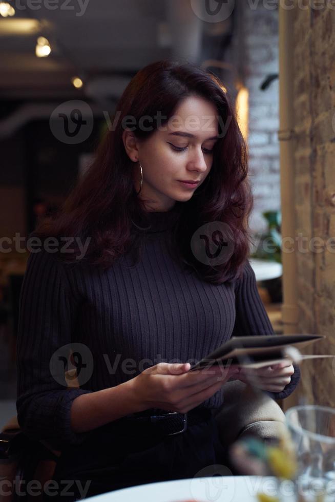 hermosa chica inteligente de moda seria y elegante sentada en un café en estilo loft en el almuerzo. listo para hacer un pedido, estudiando menú. encantadora mujer pensativa con largo cabello castaño oscuro. foto