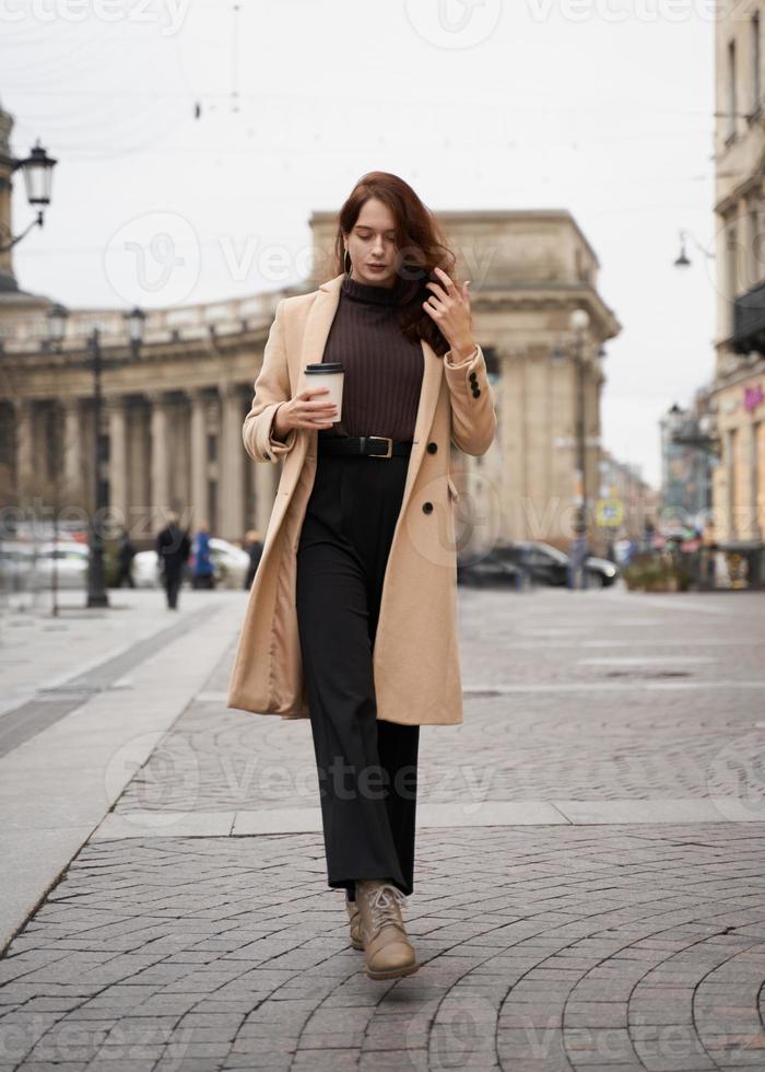 Beautiful serious stylish fashionable smart girl holding cup of coffee in hands goes walking down street of St. Petersburg in city center. Charming thoughtful woman with long dark hair photo