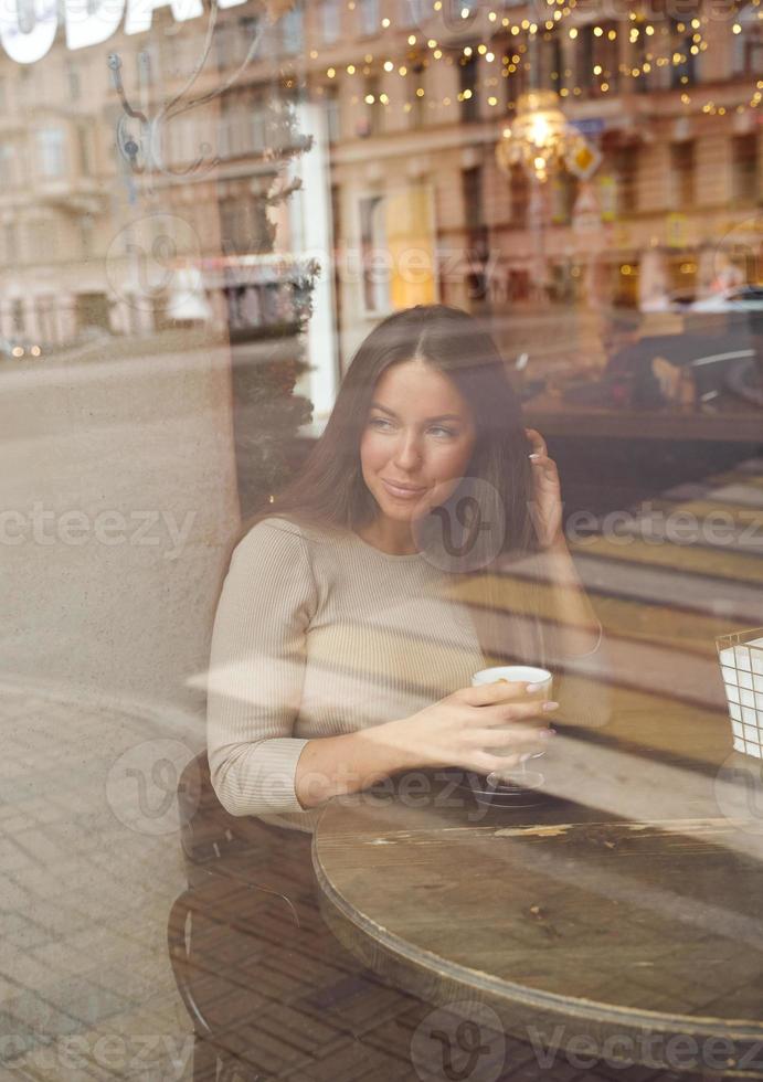A beautiful girl sits in cafe and looks out window thoughtfully. Reflection of city in window. Brunette woman with long hair smiling and drinking cappuccino coffee, vertical photo