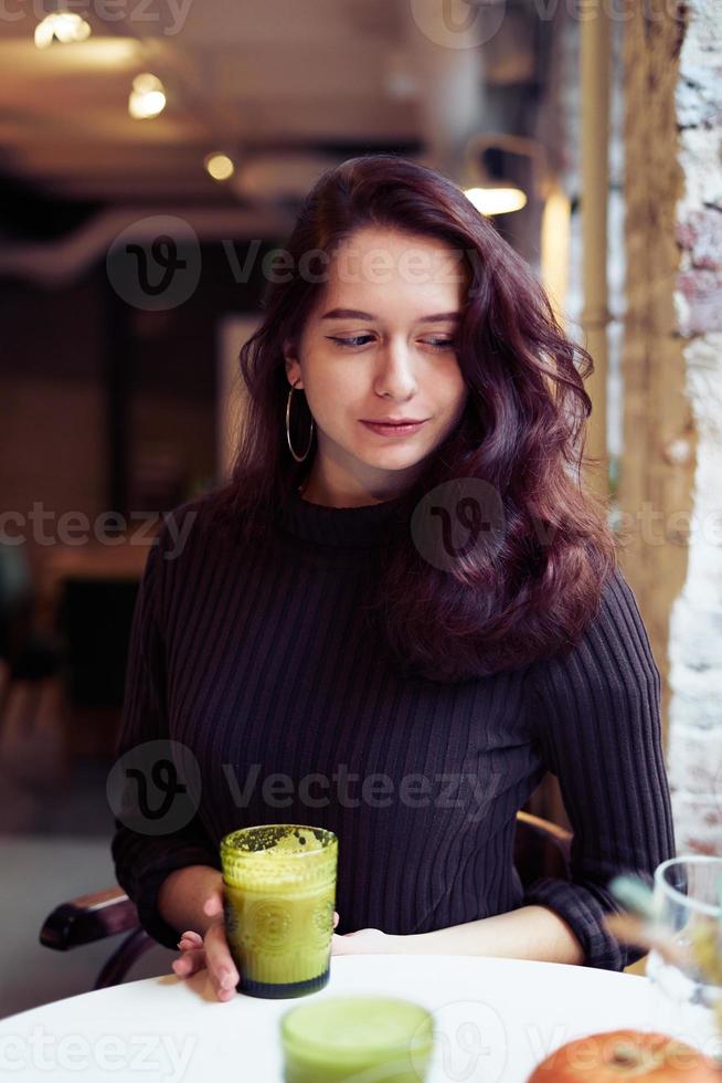 Beautiful serious stylish fashionable smart girl is sitting in cafe and drinking healthy green yellow smoothie or latte vegan. Charming thoughtful woman with long dark brown hair. photo