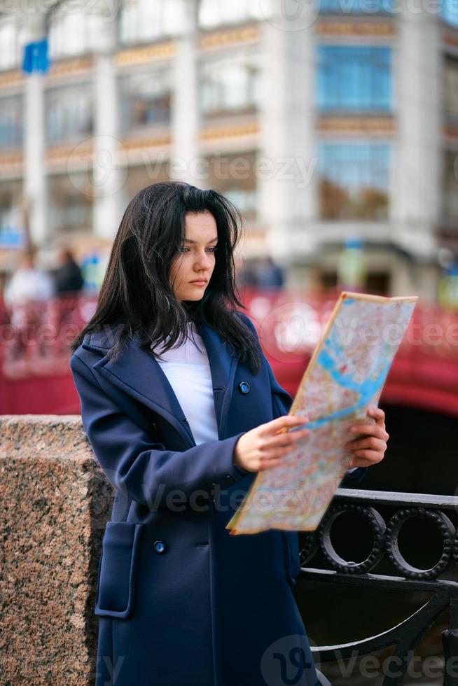 mujer joven de pie en el paseo marítimo de la gran ciudad y mirando a la guía, turista en busca de atracciones. una encantadora mujer vestida a la moda con el pelo largo y oscuro viaja por europa foto