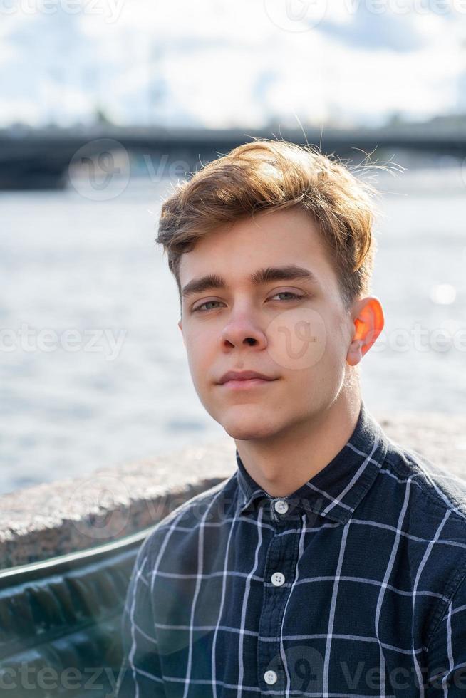 Portrait of young guy, brunette with dark hair, Caucasian. Close - up of a guy, boy, man looking at camera. Outdoor in city by water photo