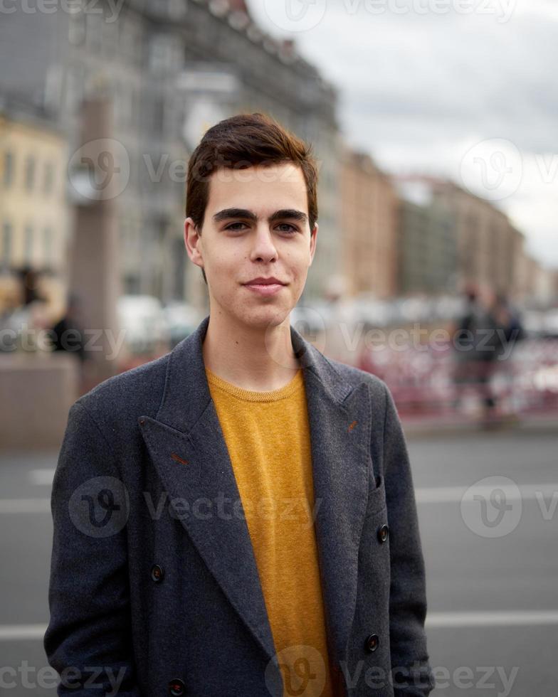 Handsome stylish fashionable man, brunette in elegant gray coat, stands on street in historical center of St. Petersburg. Young man with dark hair, thick eyebrows. photo