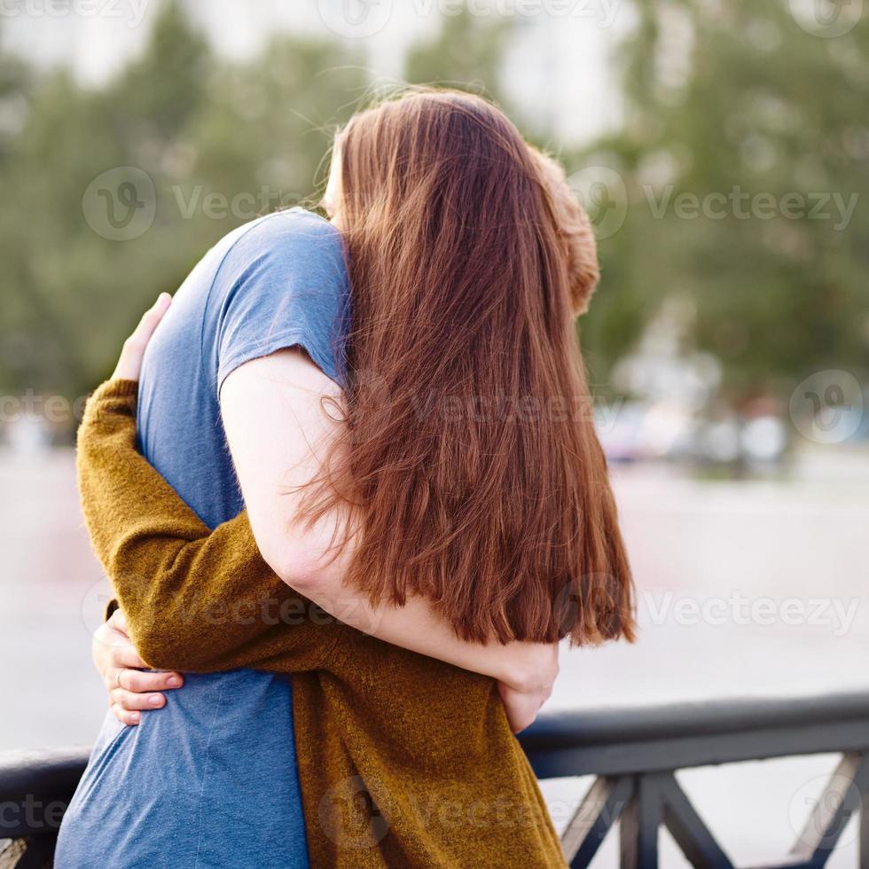 Girl with long thick dark hear embracing redhead boy on bridge, teen love at sunset photo