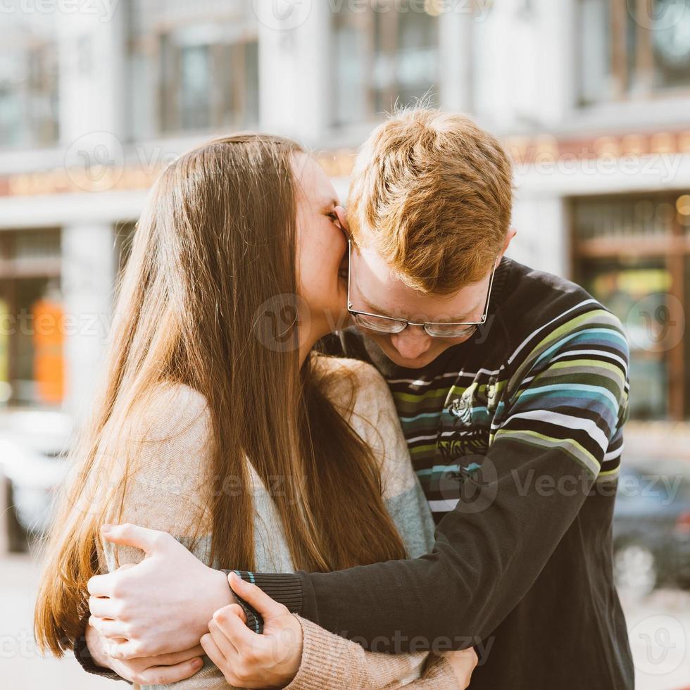 Portrait of happy couple embracing in downtown, red-haired man with glasses kisses and woman with long hair. Girl whispers in ear of guy. Concept of teenage love photo