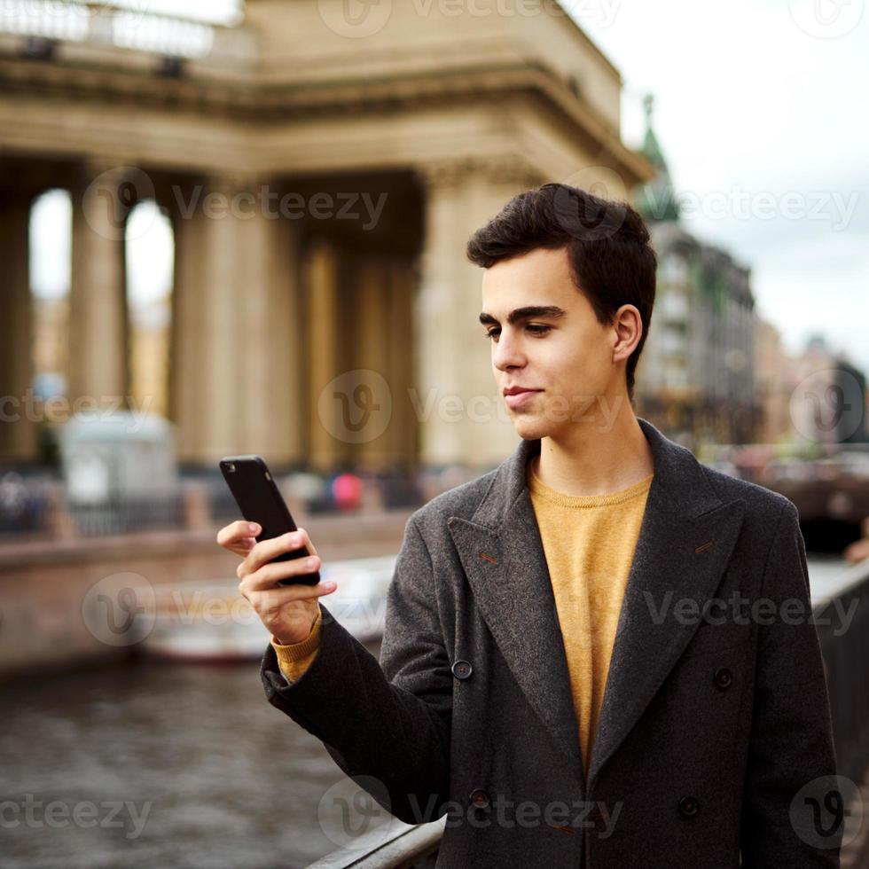 Handsome stylish fashionable man talking on phone, dialing chat message, brunette in elegant gray coat is standing on street in historical center. Young man with dark hair, thick eyebrows. photo