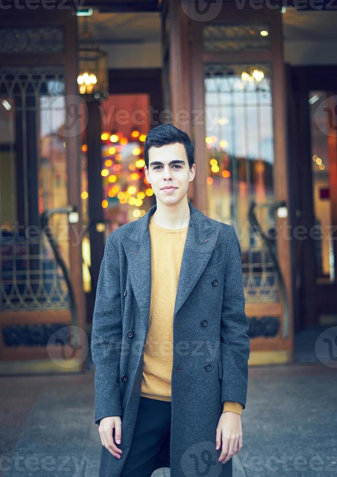 Handsome stylish fashionable man, brunette in elegant gray coat, stands on street in historical center of St. Petersburg. Young man with dark hair, thick eyebrows. photo