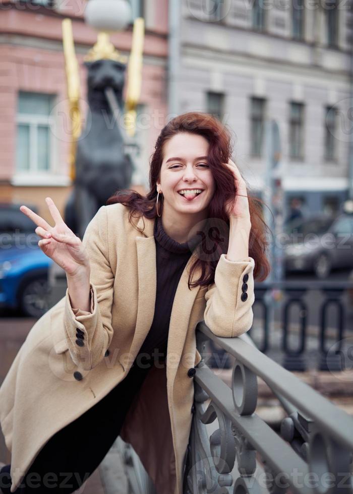 hermosa chica inteligente de moda seria y elegante de pie en el puente y sonriendo, muestra la lengua, muestra el signo de la victoria con la mano. S t. ciudad de petersburgo. encantadora mujer feliz divertida con el pelo largo y oscuro foto