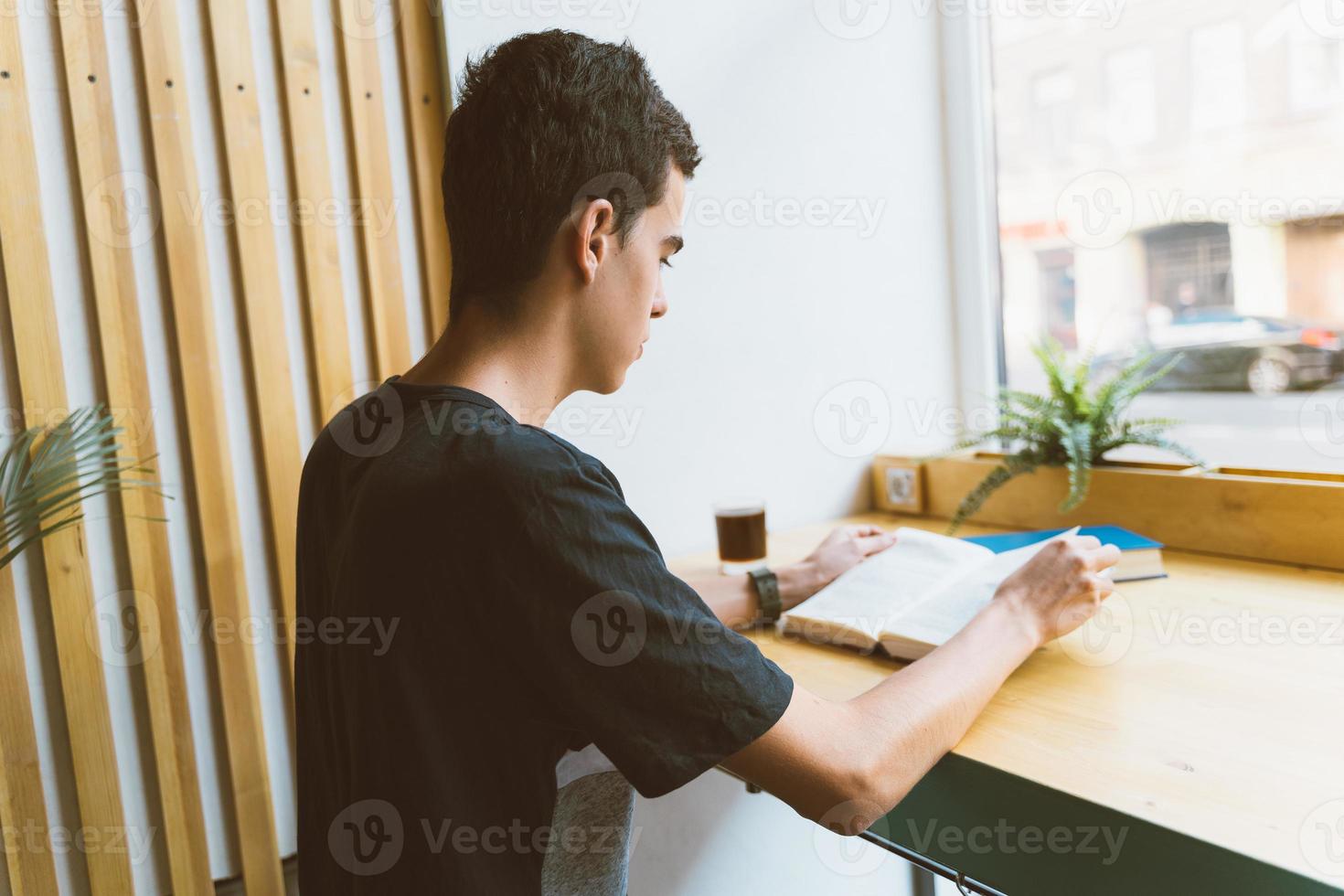 Young boy with black hair sitting by the window in the library reading a big book, teenager in casual clothes in library photo