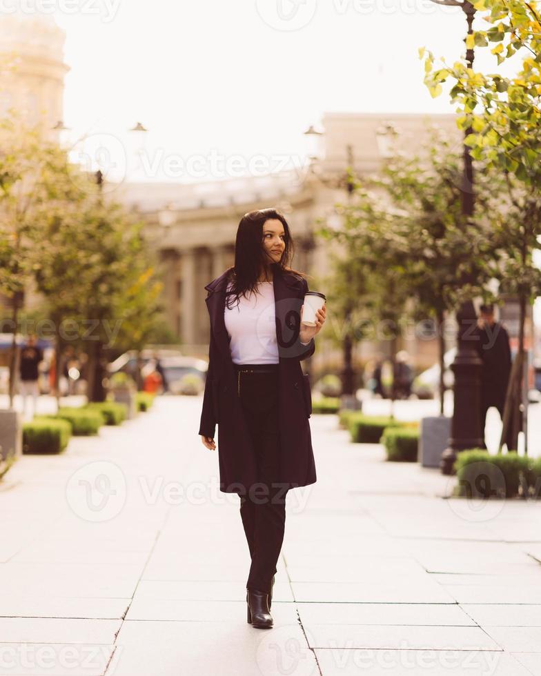 hermosa chica morena inteligente seria sosteniendo una taza de café en las manos va caminando por la calle de st. petersburgo en el centro de la ciudad. encantadora mujer pensativa con cabello largo y oscuro deambula sola foto