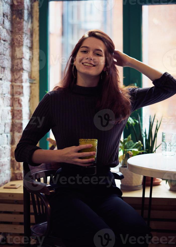 hermosa chica seria, elegante y a la moda se sienta junto a la ventana en el café, sonríe maravillosamente y bebe batido vegano saludable o café con leche. encantadora mujer pensativa con largo cabello castaño oscuro foto