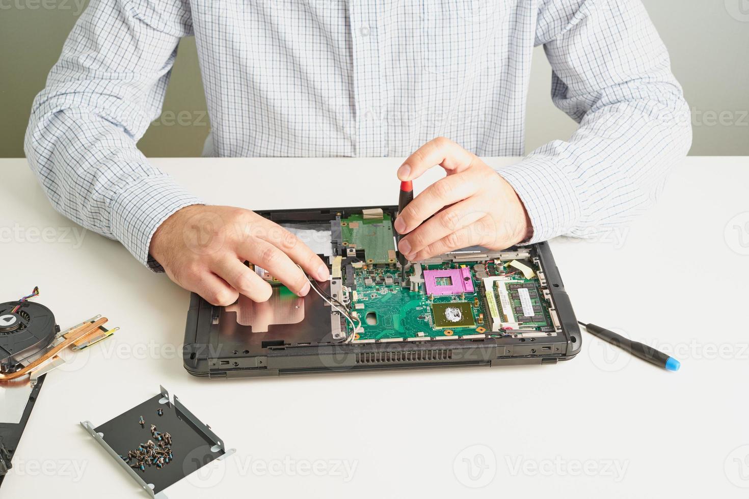 Man repairs computer. A service engineer in shirt repairs laptop, at white Desk against white wall. photo