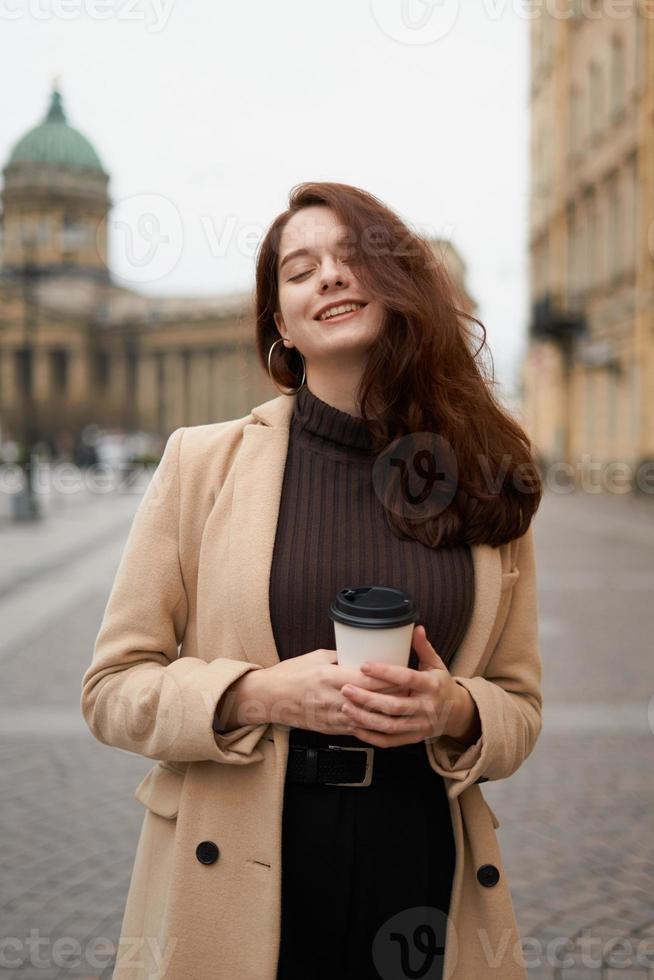 hermosa chica seria, elegante y elegante que sostiene una taza de café en las manos y sonríe, camina por la calle de st. ciudad de petersburgo. encantadora mujer pensativa con cabello largo y oscuro foto