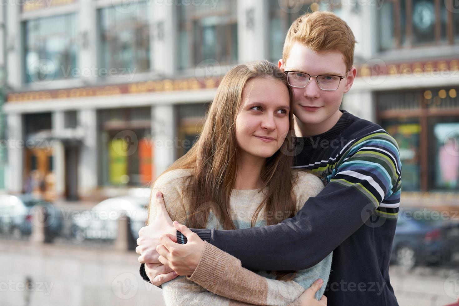 portrait of happy couple embracing in downtown, red-haired man with glasses looking straight, woman with long hair looking away photo