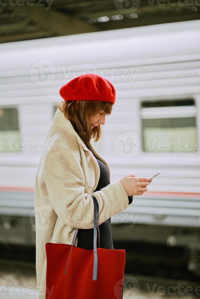 Railway station. Beautiful girl is waiting for train and looks at cell phone. Woman travels light. photo